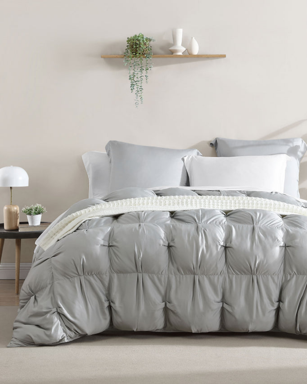 A neatly made bed with a light gray tufted comforter, white pillows, a small bedside table with a lamp and plant, and a floating shelf with decor on a beige wall.