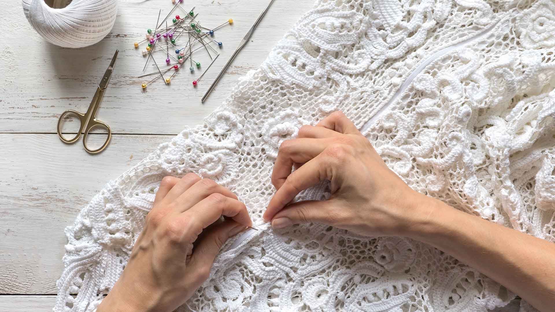 Hands sewing white lace fabric on a wooden surface, with sewing tools, pins, and scissors nearby.