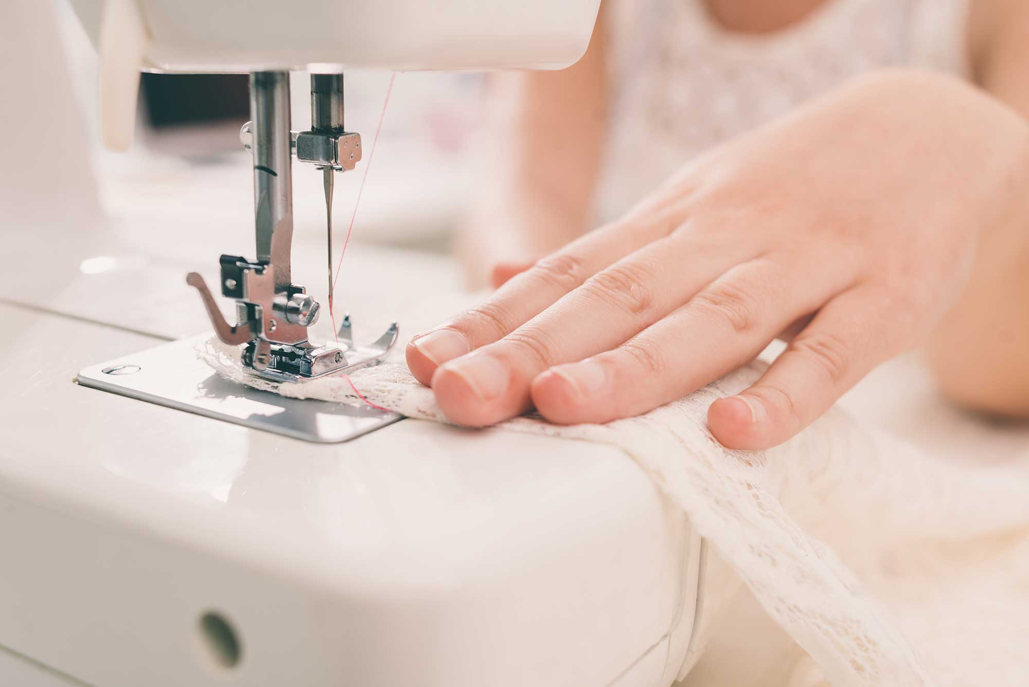 A person guides white lace fabric through a sewing machine, with their hand close to the needle.
