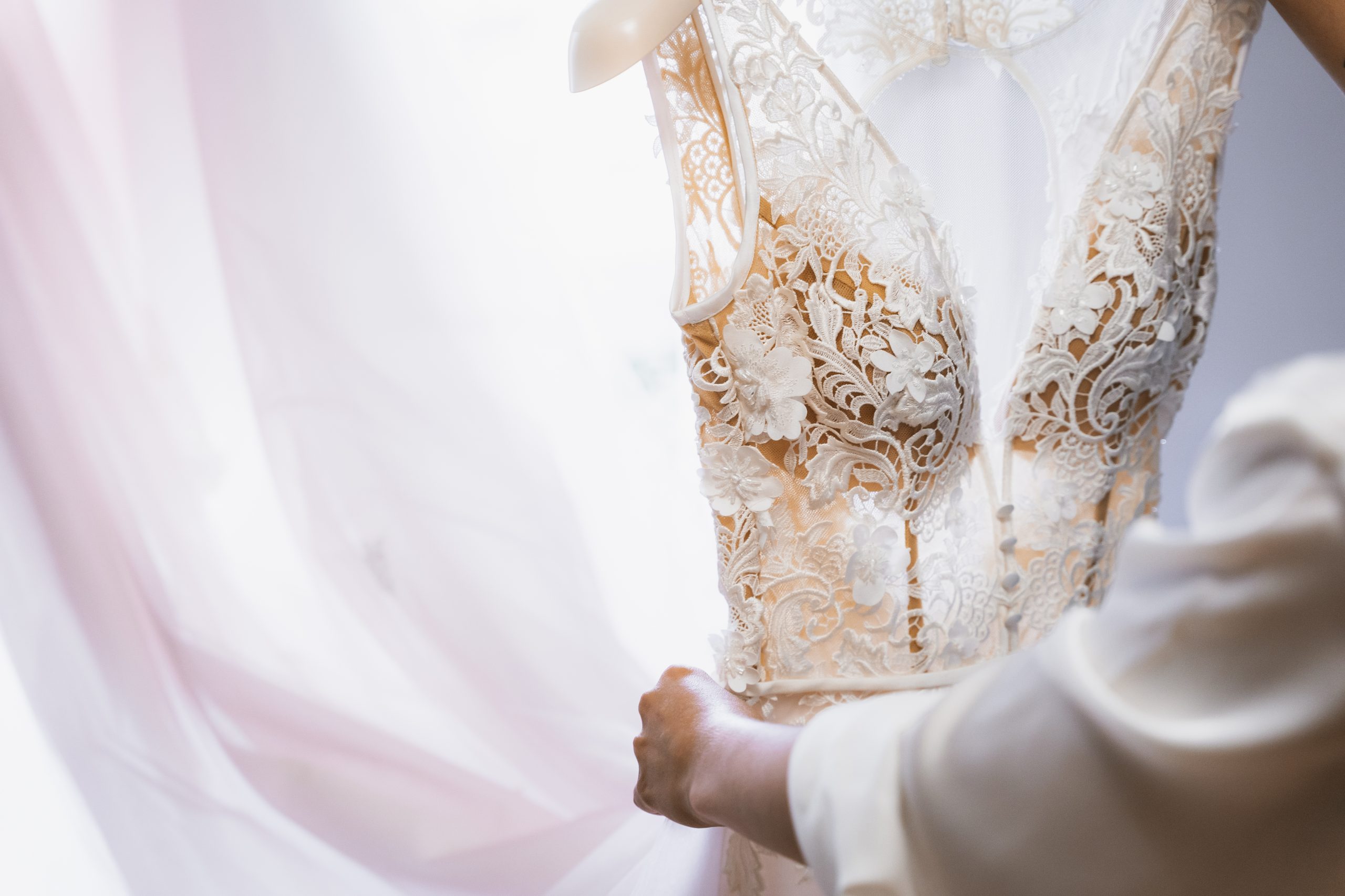 A person adjusts a detailed lace wedding dress hanging near a window with soft natural light.