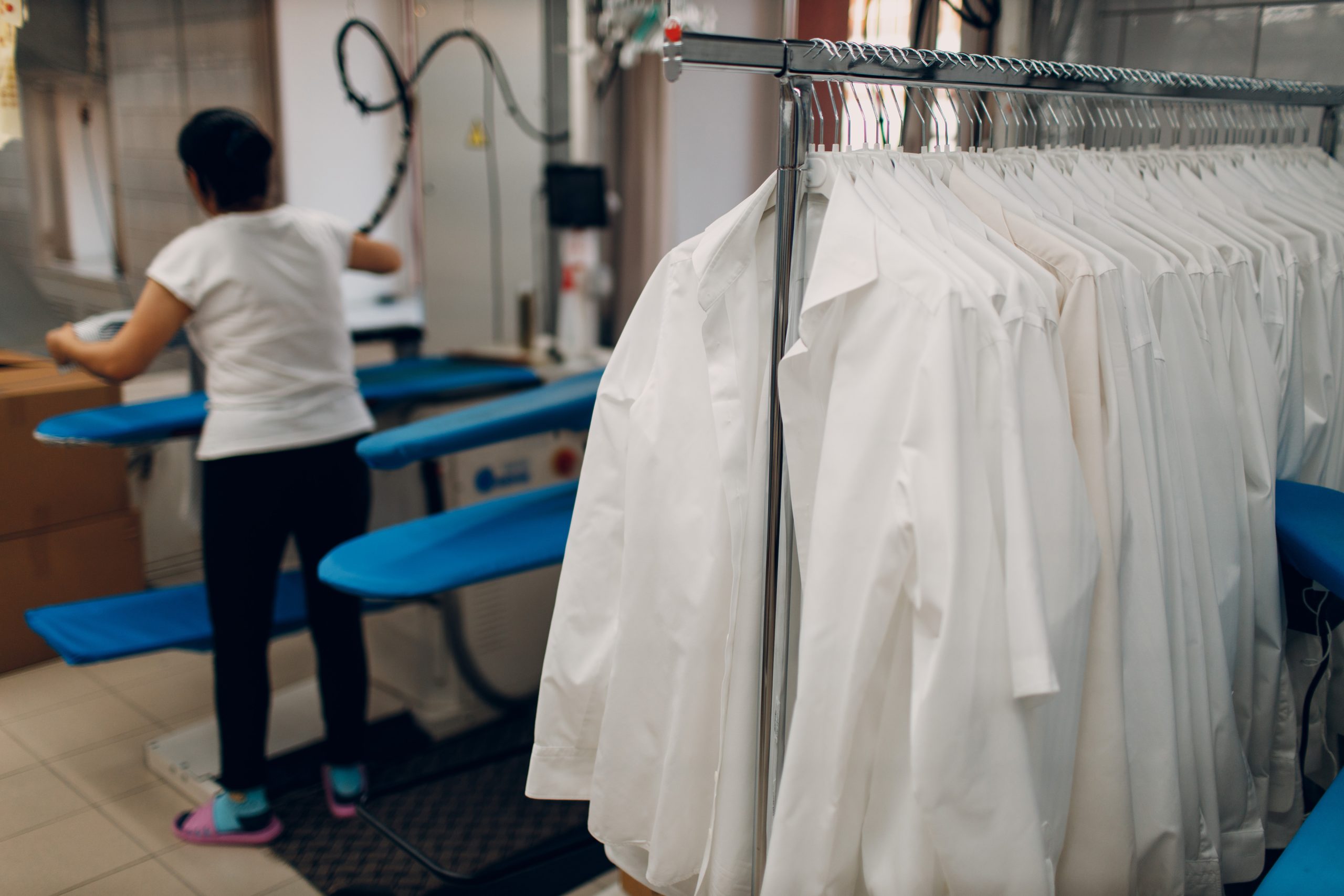 A person works in a laundry or dry cleaning facility next to a rack of freshly cleaned white lab coats hanging on hangers.