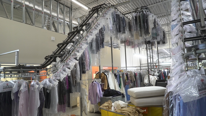 Rows of dry-cleaned clothes in plastic coverings hang on a conveyor system inside a dry cleaning facility, with various garments and laundry baskets visible.