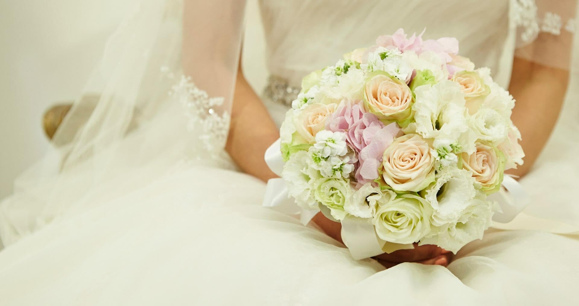 Bride in a white dress holding a bouquet of pastel-colored roses and assorted flowers, with lace details visible on her sleeves.