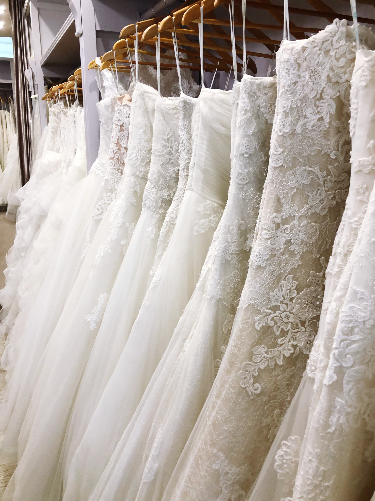 A row of white lace wedding dresses hangs on wooden hangers in a bridal shop.