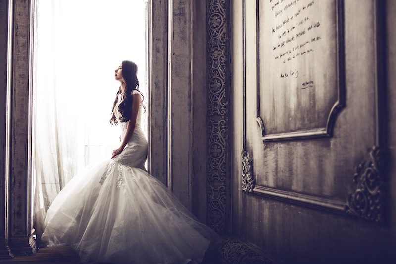 A bride in a white wedding dress sits on a windowsill, looking out, with soft light streaming through sheer curtains in an ornate, vintage room.