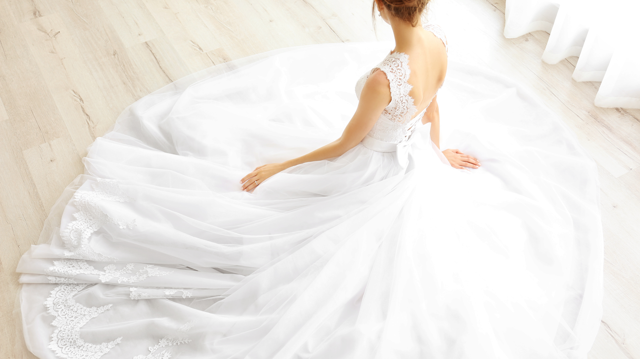 A woman in a white lace wedding dress sits on a light wood floor, viewed from above, with the dress spread out in a circle around her.
