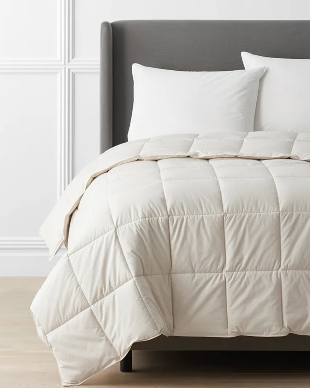 A neatly made bed with a light-colored, quilted comforter and two white pillows, featuring a gray upholstered headboard against a white paneled wall.